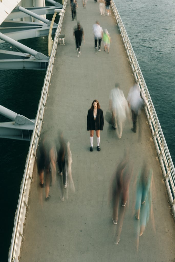 A woman stands still on a bustling footbridge, capturing a moment of solitude amidst motion.