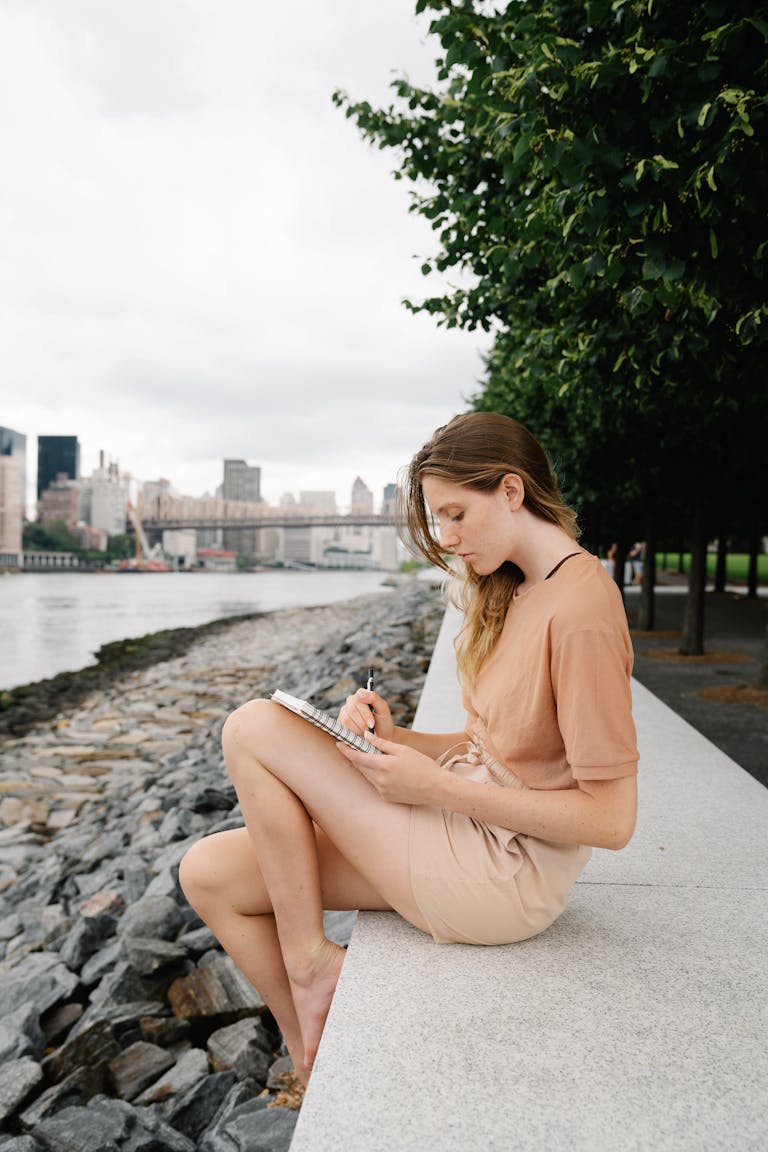 A young woman sits by a city waterfront, writing in a notebook on a cloudy day.