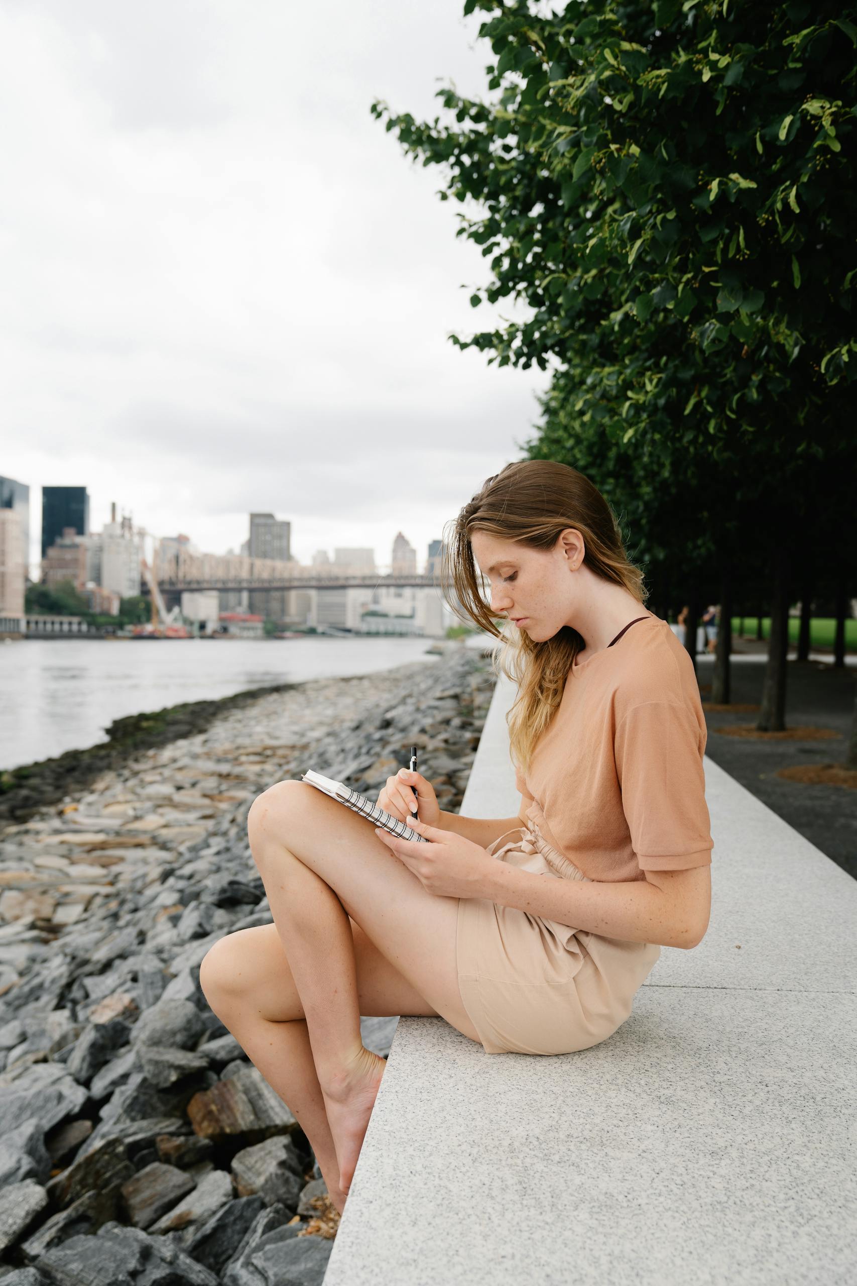 A young woman sits by a city waterfront, writing in a notebook on a cloudy day.