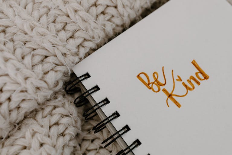 Handwritten 'Be Kind' affirmation on a notebook with a warm knitted blanket background.