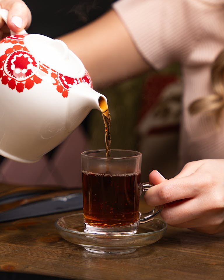 A woman pours tea from a teapot into a glass cup on a wooden table. Warm and cozy atmosphere.
