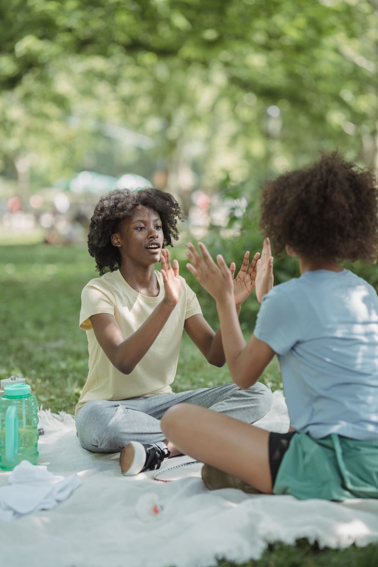 Two teenage girls enjoy a playful moment of patty cake during a picnic in a sunny park.